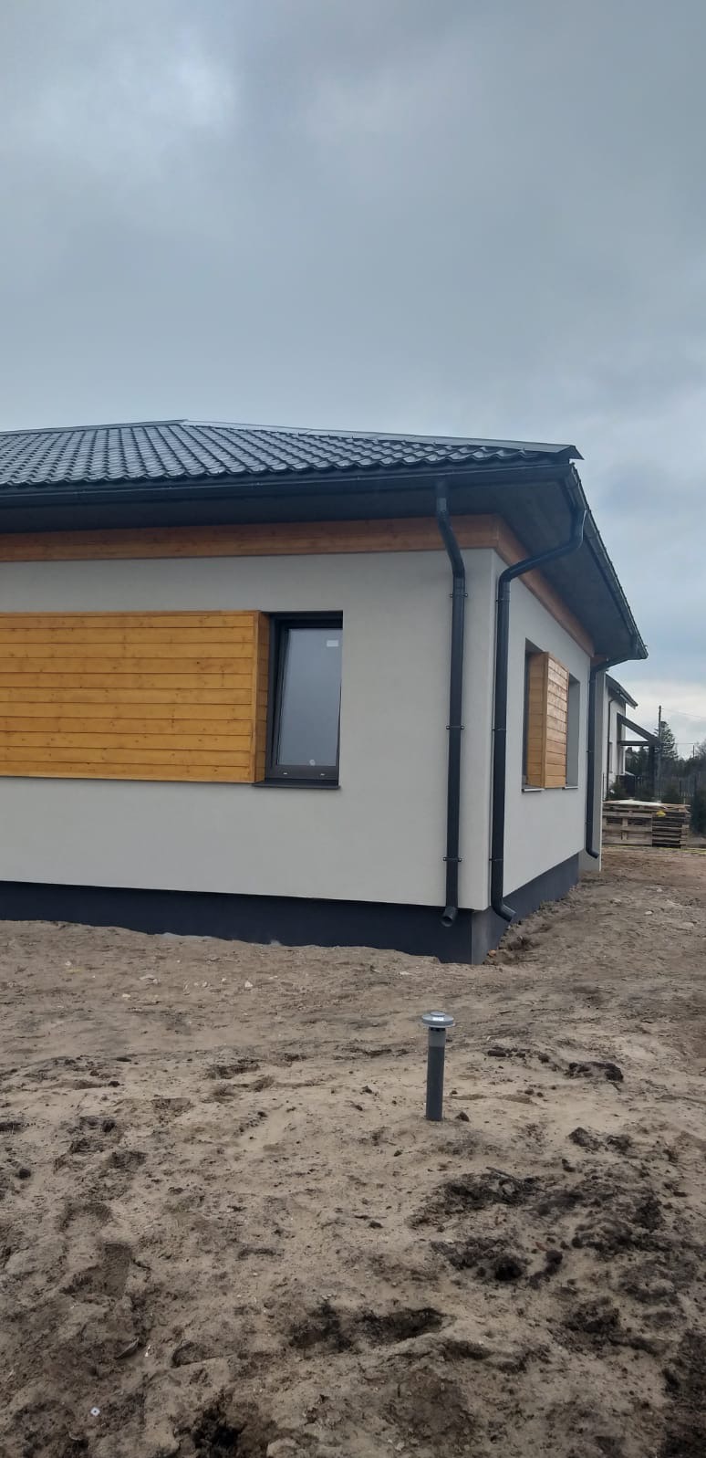 Nowoczesny dom jednorodzinny z szarą elewacją, drewnianymi panelami dekoracyjnymi pod oknami i ciemną dachówką, widoczny z zewnątrz na etapie wykończenia terenu.