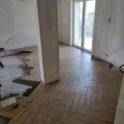 Łukasz Bączkowski - Układanie parkietu w jodełkę w pomieszczeniu z surowymi ścianami i kolumną, widoczne narzędzia i niedokończony fragment podłogi.