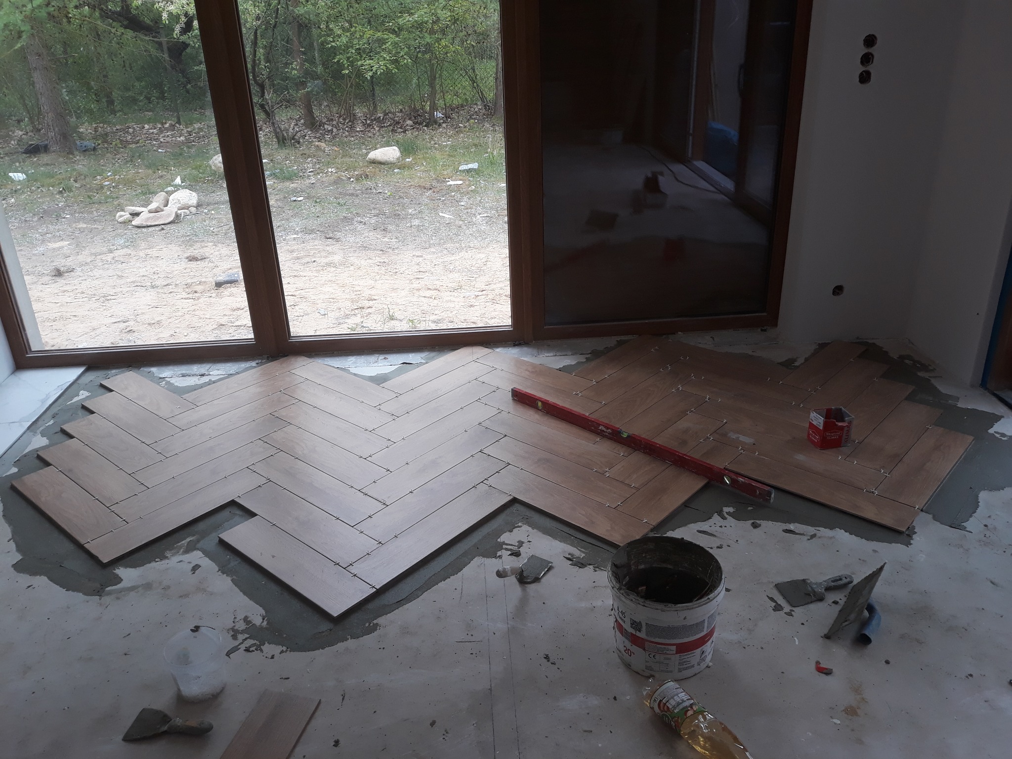 Układanie drewnopodobnych płytek podłogowych w jodełkę, widoczny poziomica, wiadro z klejem i narzędzia glazurnicze na surowym podłożu, duże okno z widokiem na zewnątrz.