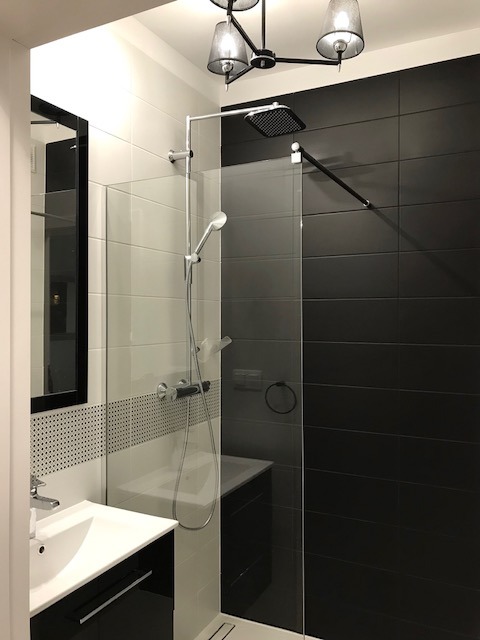 Nowoczesna łazienka z czarno-białymi płytkami, szklaną kabiną prysznicową typu walk-in i czarną szafką pod umywalką, oświetlona designerską lampą sufitową.