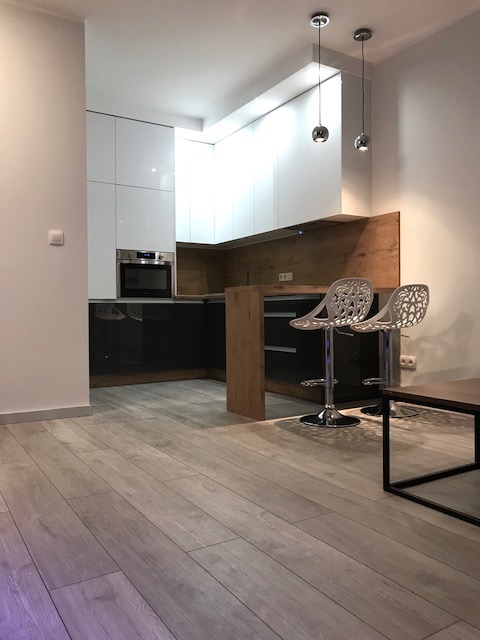 Nowoczesna kuchnia z białymi szafkami górnymi i ciemnymi dolnymi, drewnianym blatem i dwoma hokerami o ażurowych oparciach, oświetlona wiszącymi lampami kulami.