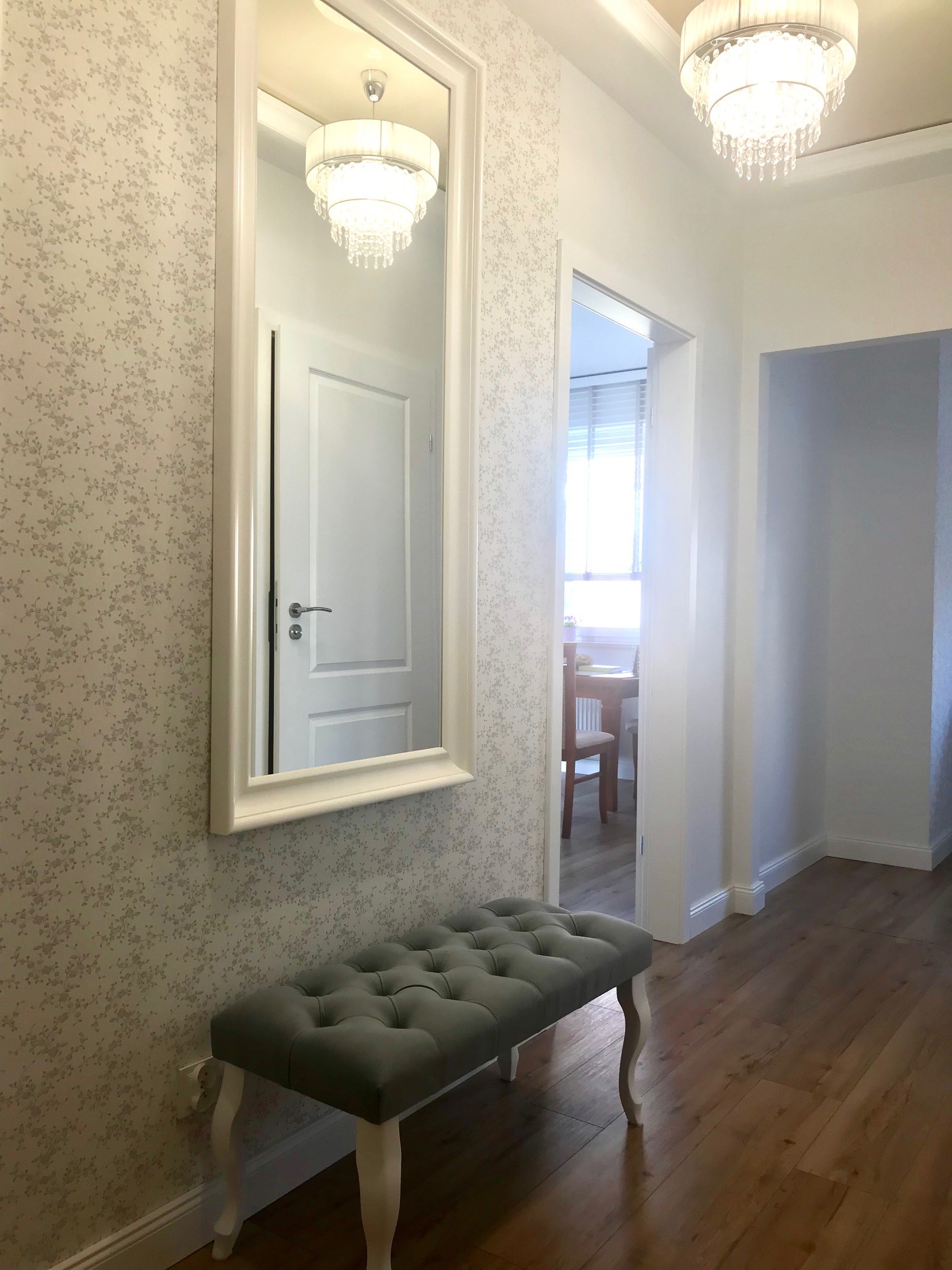 Jasny korytarz z tapetą w drobne kwiaty, lustrem w białej ramie, szarą ławką z pikowanym siedziskiem i widokiem na pokój z drewnianym stołem i krzesłami.