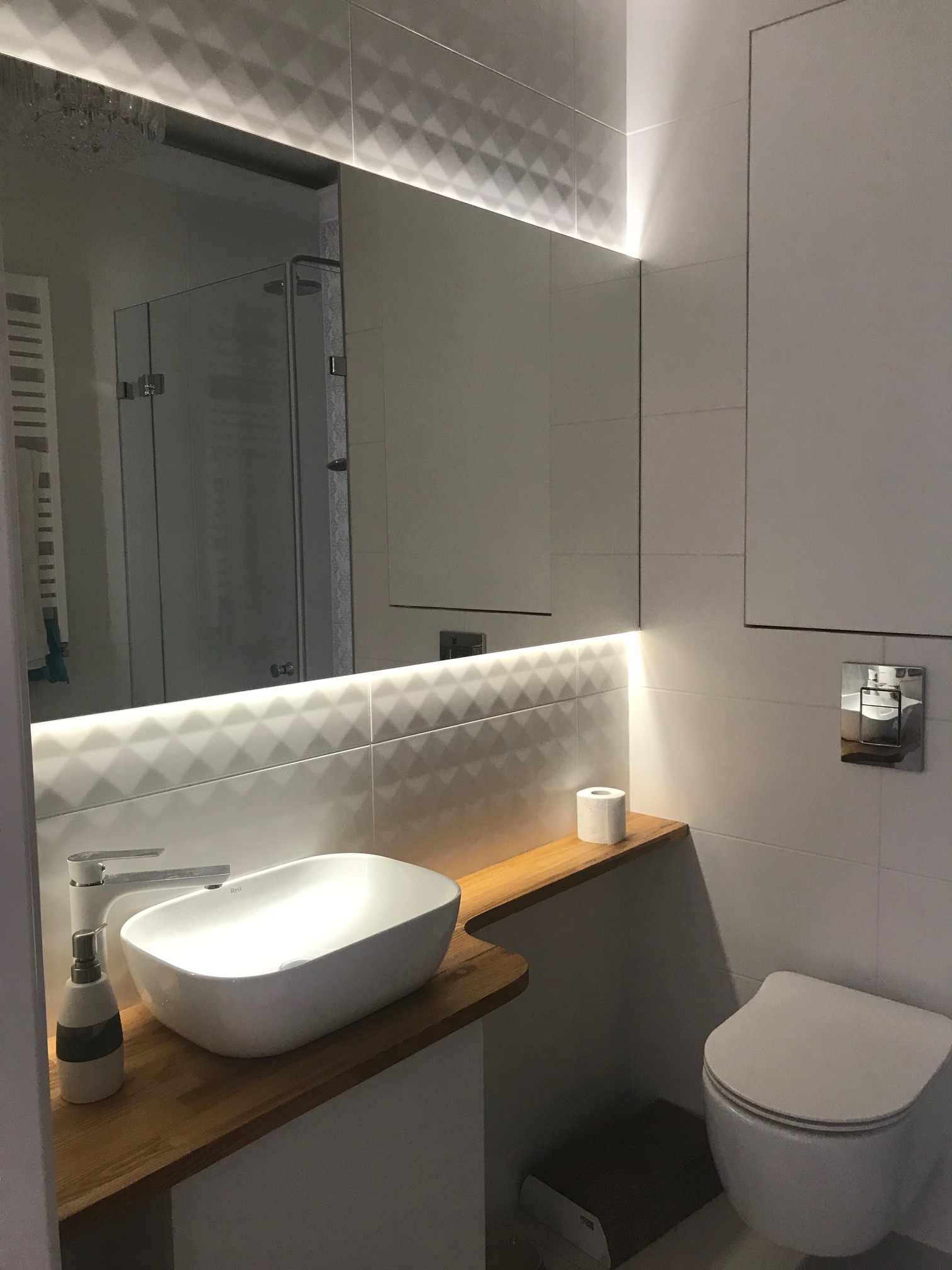 Nowoczesna łazienka w jasnych kolorach z umywalką nablatową, drewnianym blatem, podświetlanym lustrem i wiszącą toaletą.