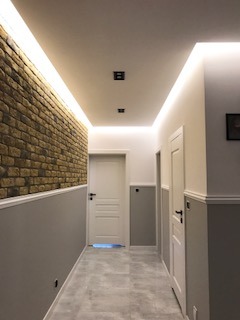 Korytarz z oświetleniem LED podsufitowym, ściana z cegły dekoracyjnej, dolna część ścian pomalowana na szaro, białe drzwi z czarnymi klamkami, szara podłoga z płytek.