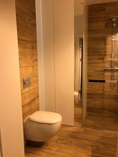 Nowoczesna łazienka z toaletą podwieszaną, wykończona panelami imitującymi drewno na ścianach i podłodze, kabina prysznicowa ze szklaną ścianą, minimalistyczna armatura.