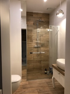 Nowoczesna łazienka z kabiną prysznicową typu walk-in, wykończona płytkami imitującymi drewno na ścianie i podłodze. Widoczna umywalka nablatowa na stylizowanej szafce i szklana kula jako lampa.