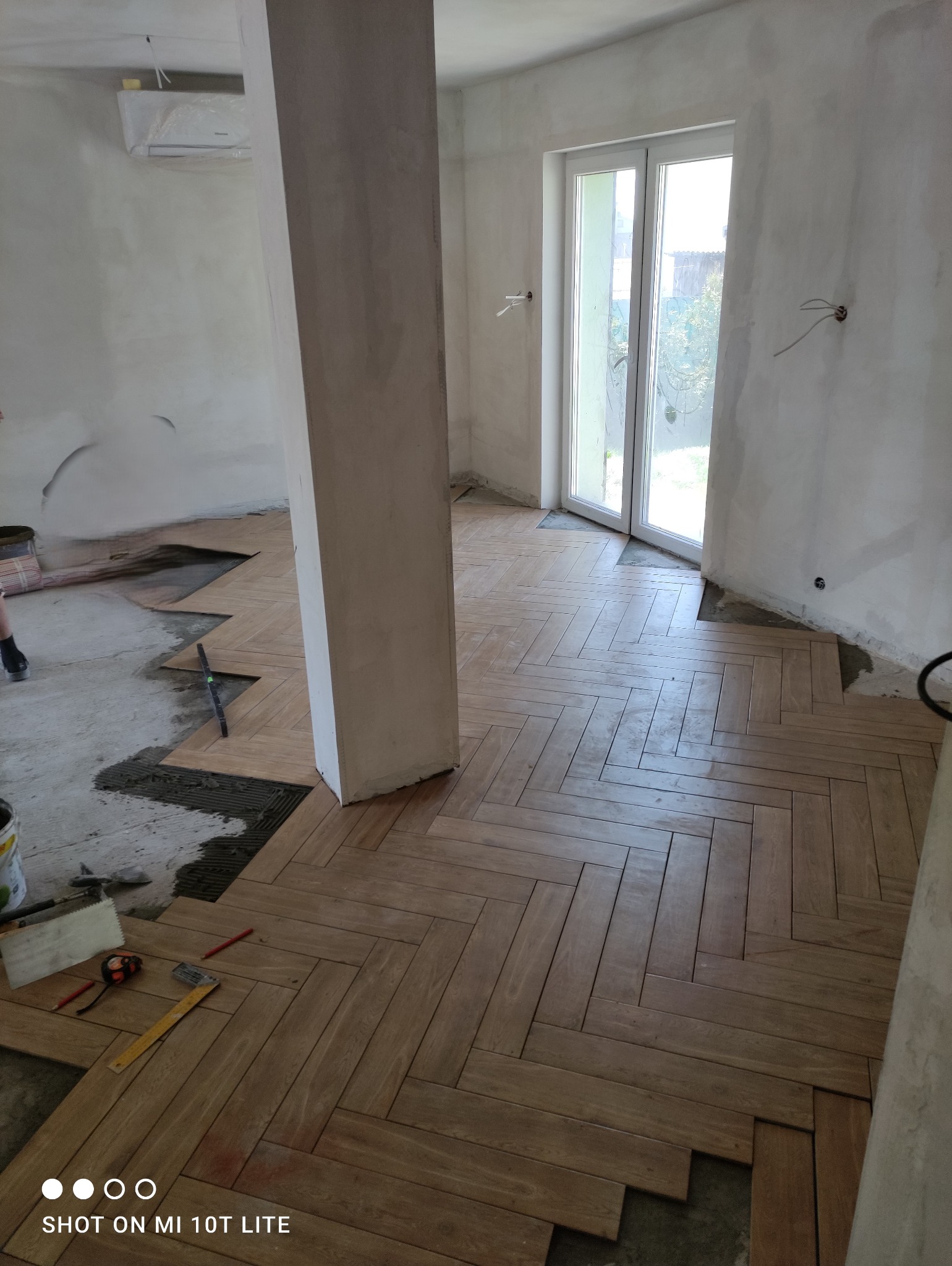 Układanie parkietu w jodełkę w pomieszczeniu z surowymi ścianami i kolumną, widoczne narzędzia i niedokończony fragment podłogi.