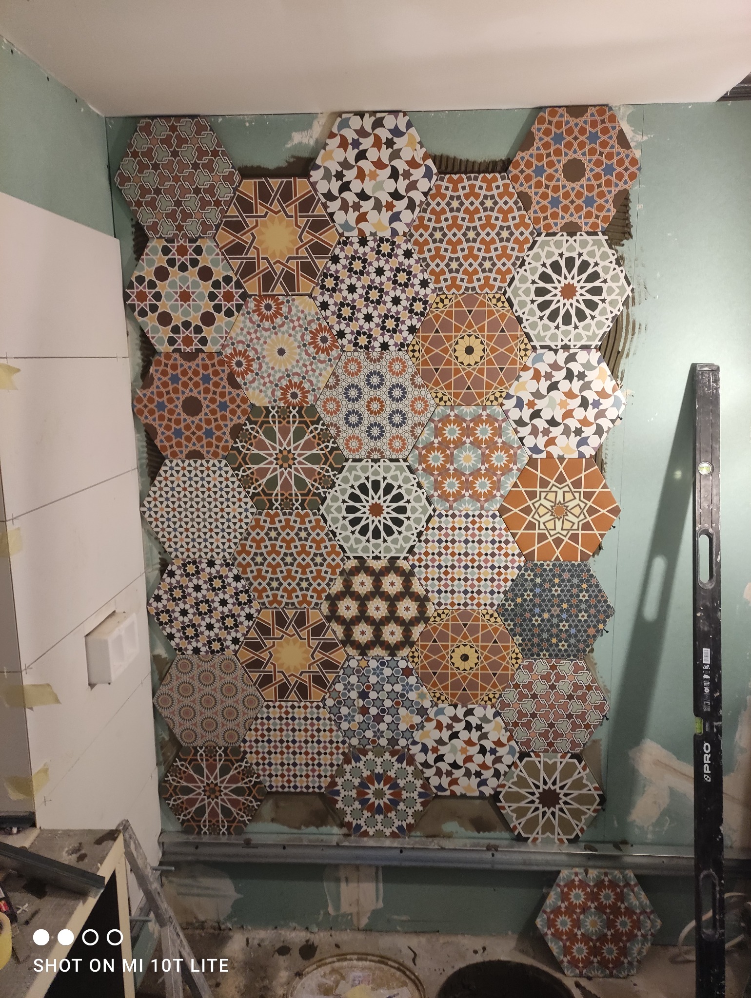 Montaż dekoracyjnych, heksagonalnych płytek ściennych z motywami geometrycznymi na ścianie w trakcie prac wykończeniowych, widoczna poziomica i fragment instalacji sanitarnej.
