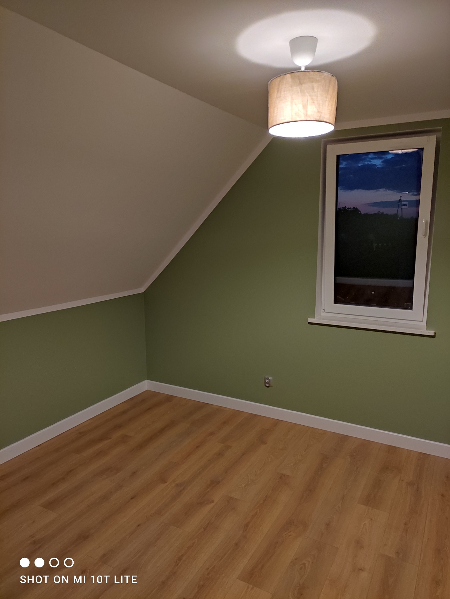 Pokój na poddaszu po malowaniu ścian na zielono, z drewnianą podłogą, białymi listwami przypodłogowymi i widokiem na nocne niebo za oknem.