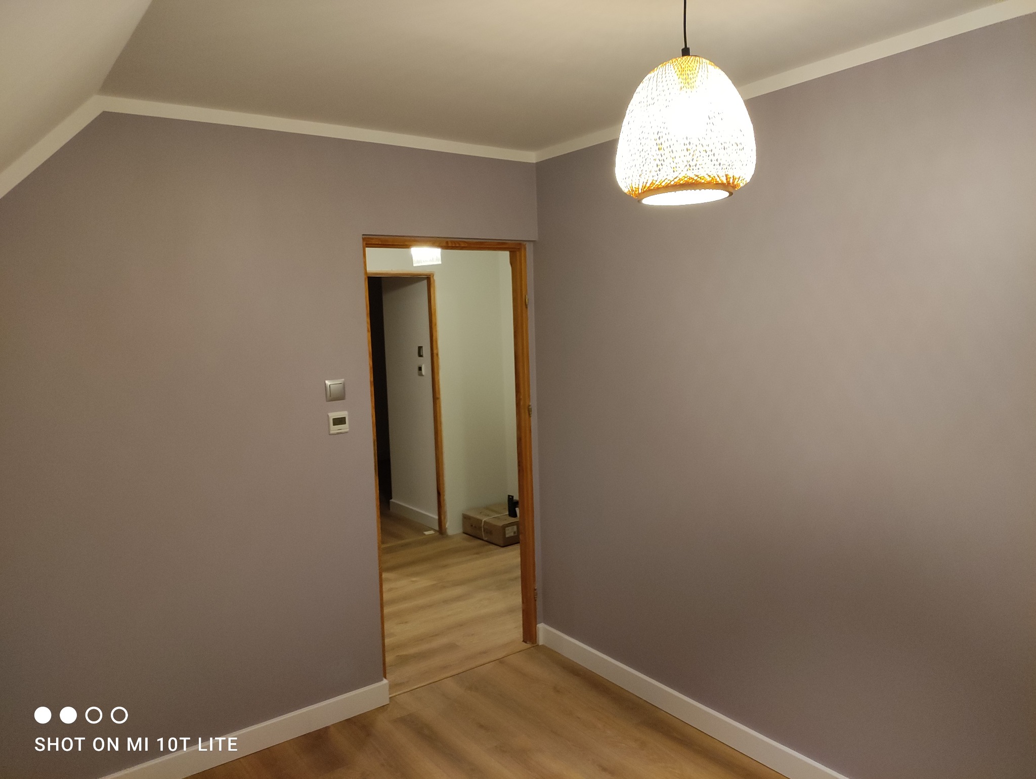 Pomieszczenie po remoncie: ściany pomalowane na szaro, białe listwy przypodłogowe, drewniana podłoga, widoczny fragment sufitu ze zwisającą lampą z abażurem, widok przez otwarte drzwi do kolejnego...