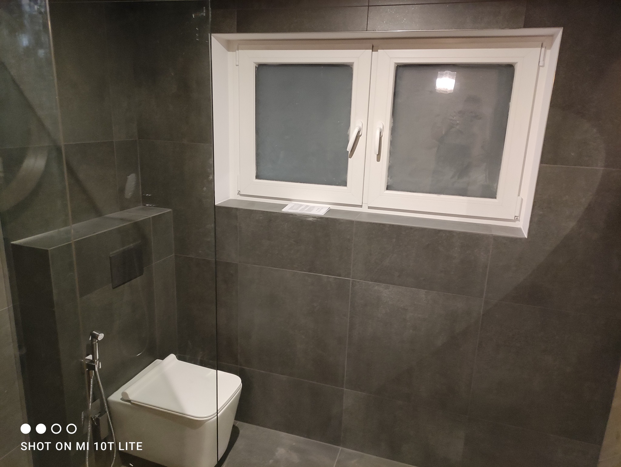 Nowoczesna łazienka z szarymi płytkami, podwieszaną toaletą, szklaną kabiną prysznicową i białym oknem z parapetem.