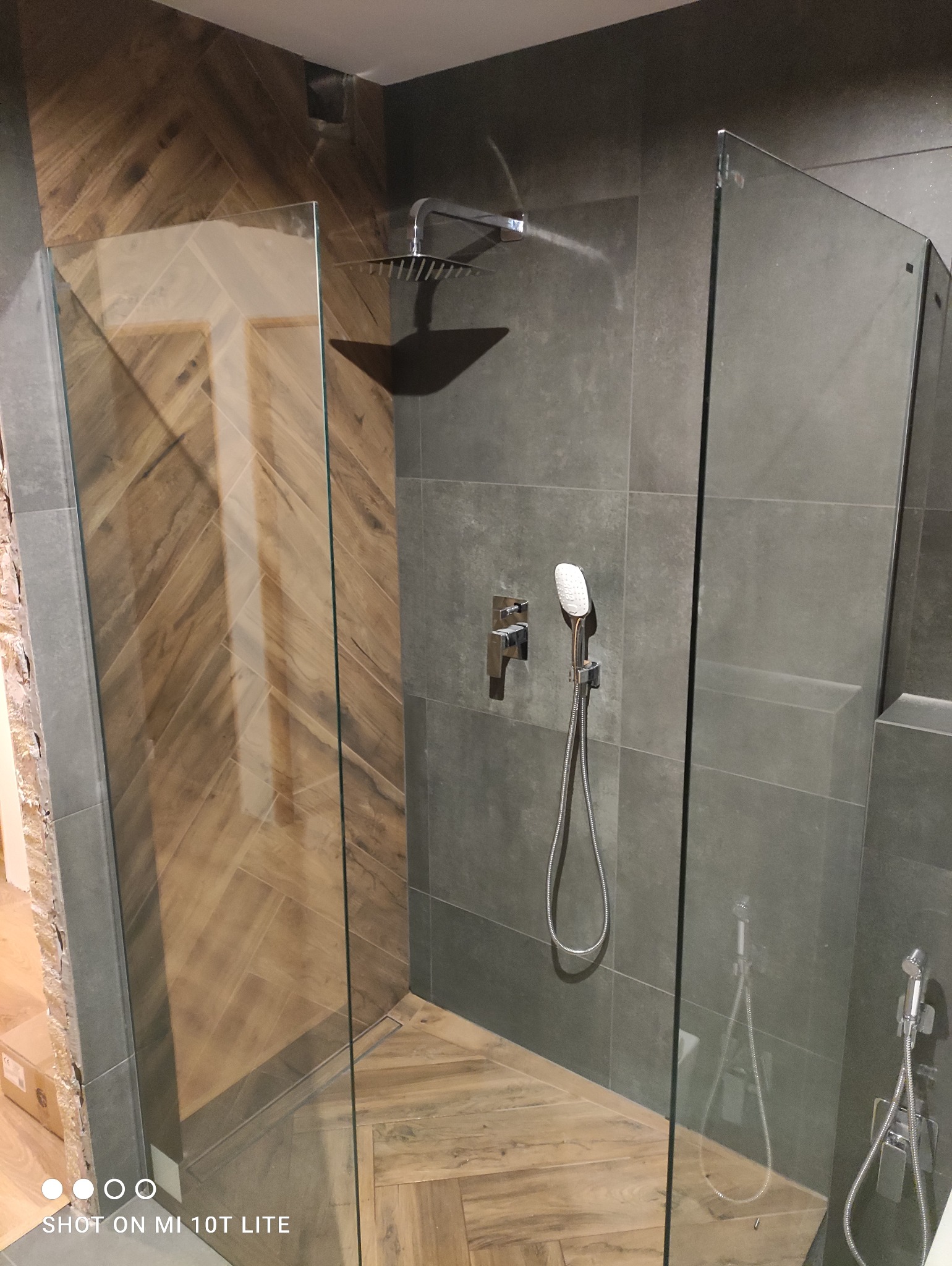 Nowoczesna kabina prysznicowa z deszczownicą i szklanymi ściankami, wykończona szarą płytką i drewnopodobną podłogą ułożoną w jodełkę.