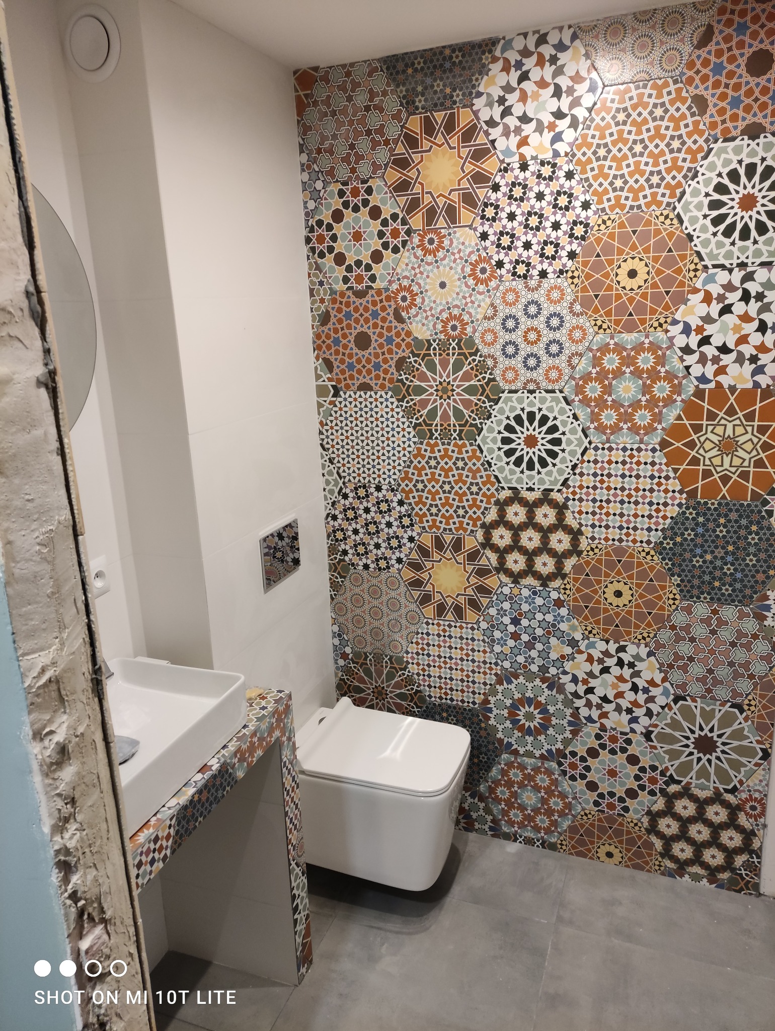 Wykończona łazienka z mozaikową ścianą i umywalką, widoczna biała toaleta podwieszana i szara podłoga, ściana w heksagonalne płytki z motywami geometrycznymi w odcieniach brązu, beżu i niebieskiego.