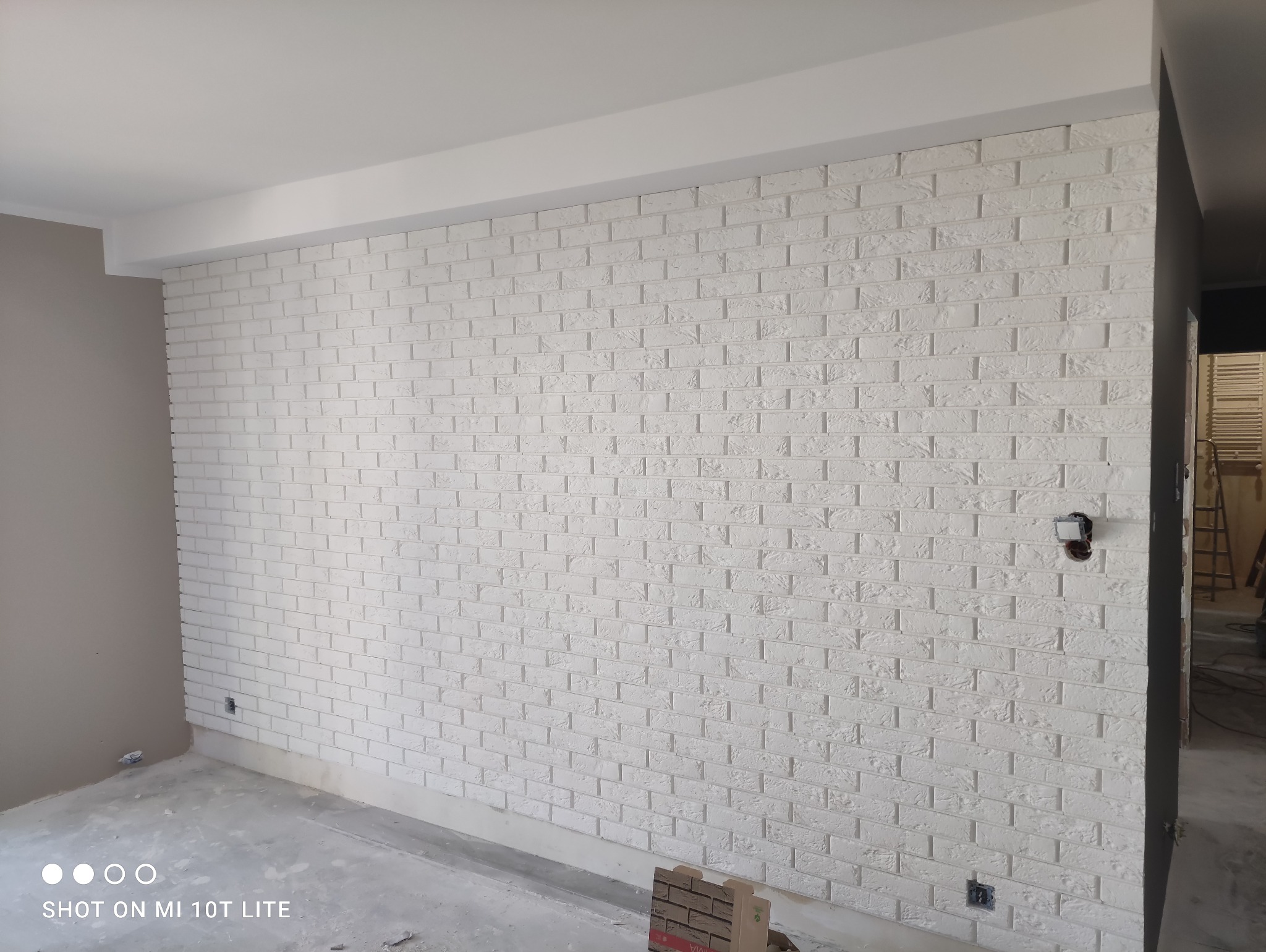 Ściana wykończona białą cegłą dekoracyjną w nowym wnętrzu, widoczny fragment podłogi w trakcie prac oraz puszka elektryczna bez osprzętu.