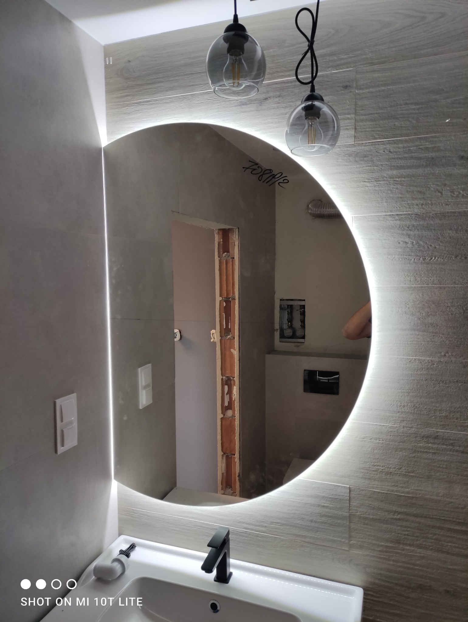 Okrągłe lustro z podświetleniem LED w łazience, odbijające fragment ściany z widoczną instalacją hydrauliczną i fragmentem surowej ściany z cegłami, zawieszone nad białą umywalką z czarną baterią...