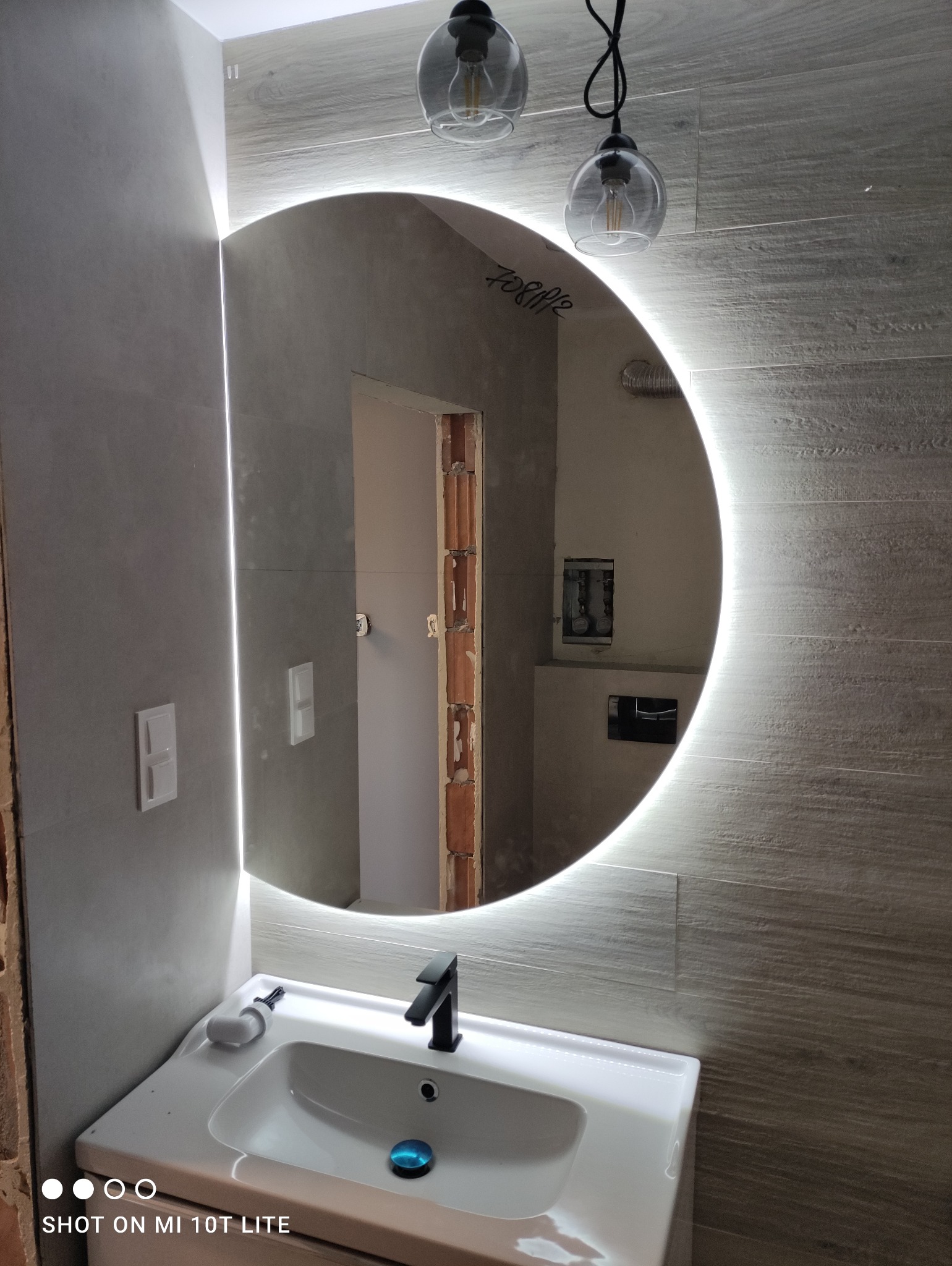 Łazienka w trakcie wykańczania z okrągłym lustrem podświetlanym LED, widoczne instalacje, biała umywalka i drewnopodobne płytki na ścianie.