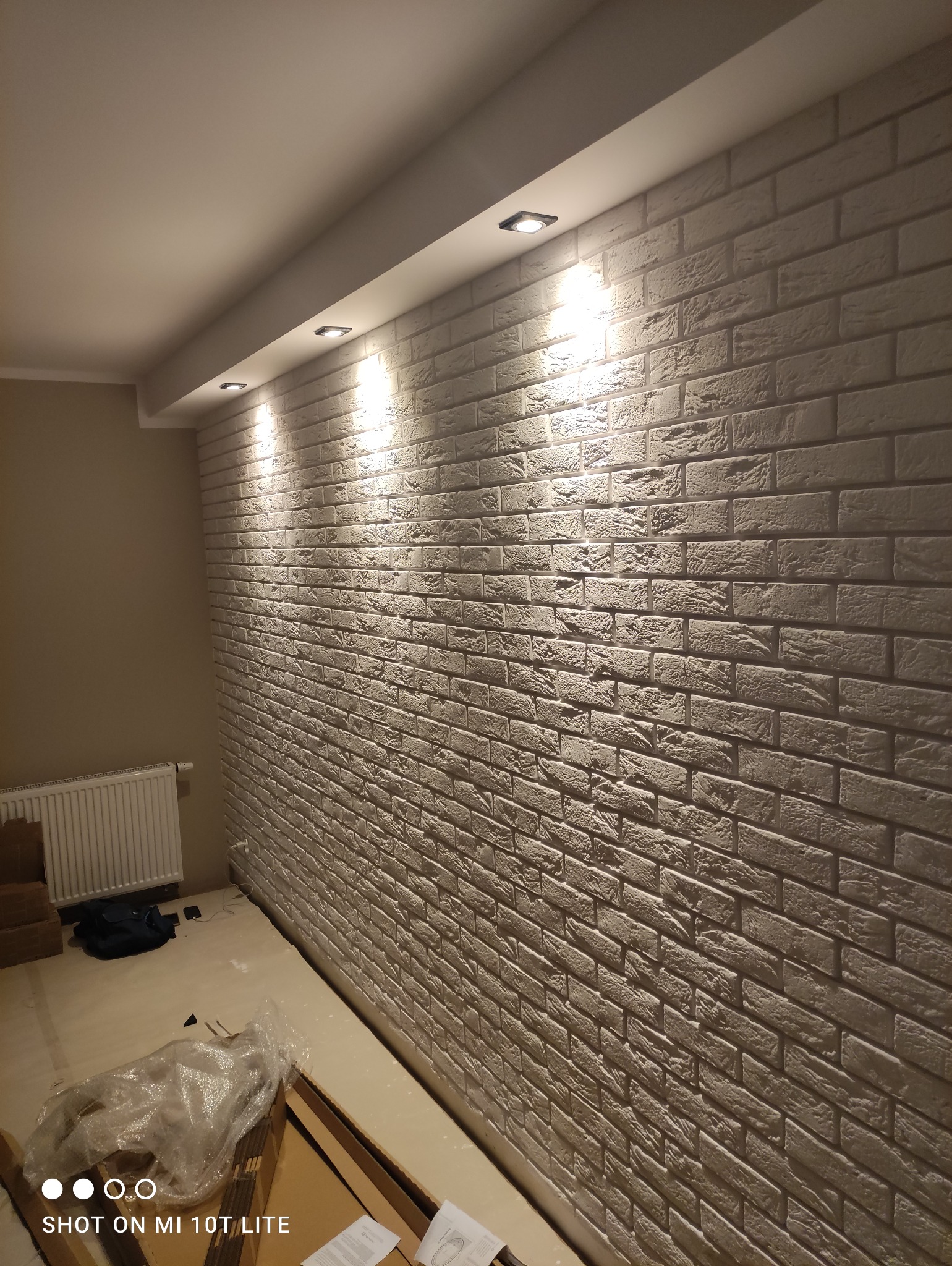 Ściana wykończona dekoracyjną cegłą w odcieniach szarości, oświetlona trzema punktowymi lampami sufitowymi, widoczny grzejnik i częściowo rozpakowane materiały budowlane na podłodze.