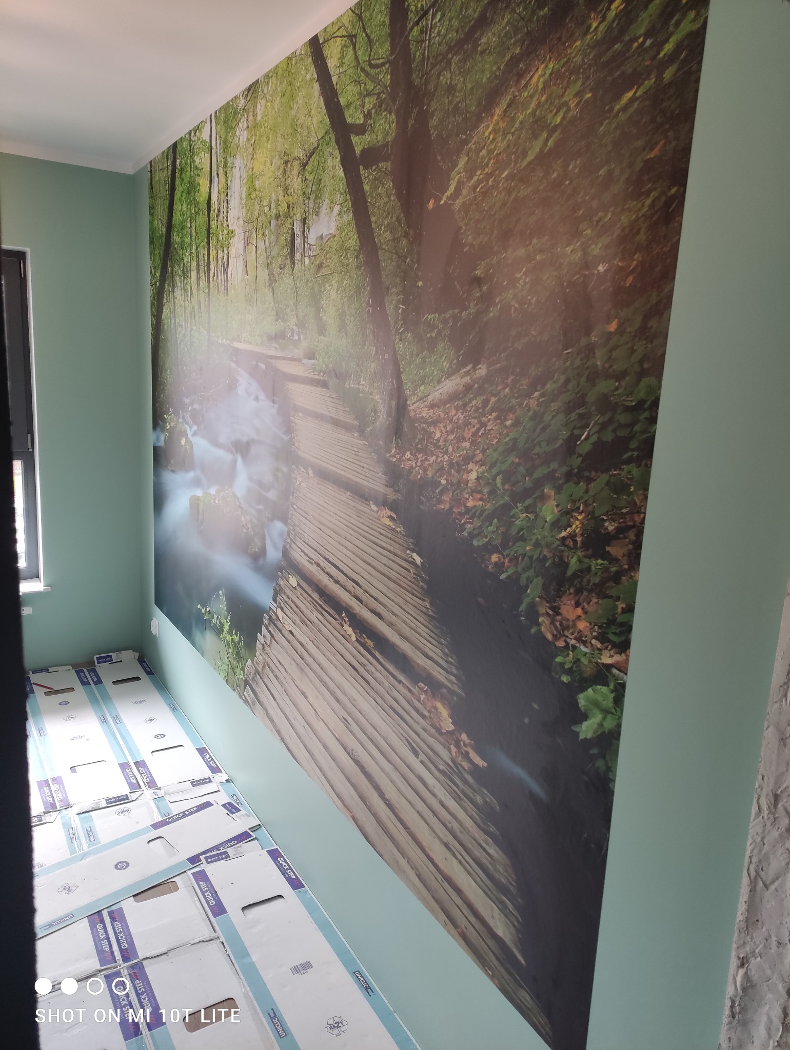 Fototapeta z motywem leśnej ścieżki nad wodospadem, przy podłodze leżą kartony z panelami podłogowymi Quick-Step, ściany pomalowane na jasny zielony kolor.