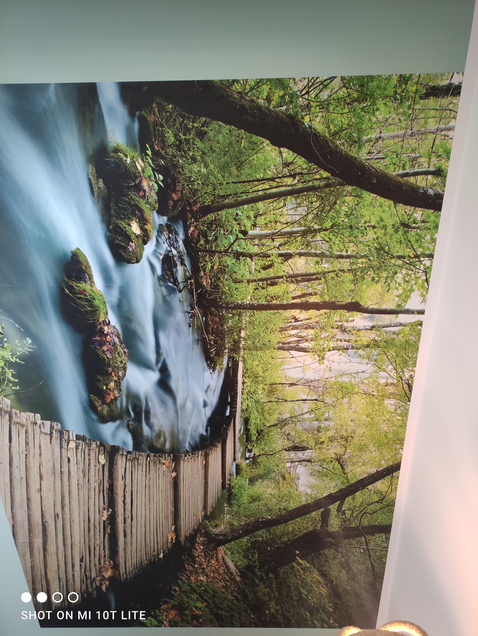 Fototapeta przedstawiająca drewniany most nad rwącym potokiem w lesie, z długim czasem naświetlania, ukazana na ścianie we wnętrzu.