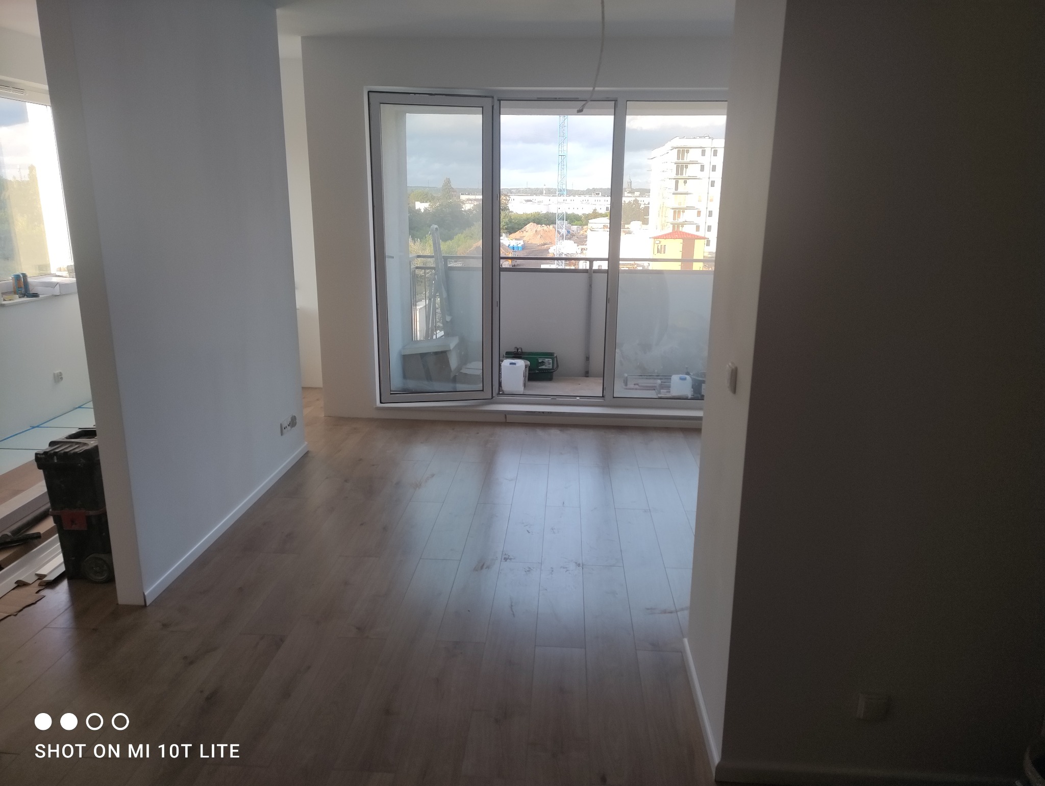 Pusty pokój z nową drewnianą podłogą, świeżo pomalowanymi białymi ścianami i otwartym balkonem z widokiem na miasto w tle, narzędzia i materiały budowlane w rogu pokoju sugerują trwające prace.