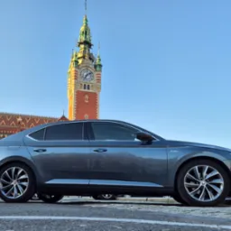 Szary samochód Skoda Superb zaparkowany na tle budynku z wieżą zegarową.