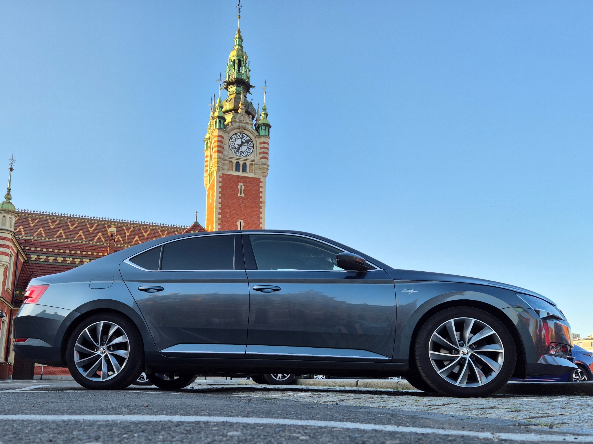 Szary samochód Skoda Superb zaparkowany na tle budynku z wieżą zegarową.