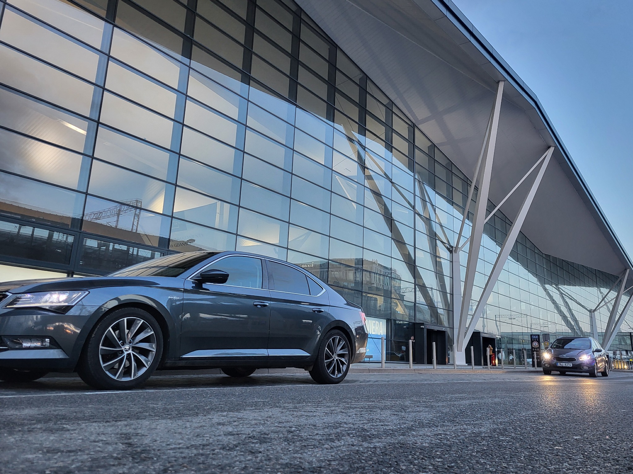 Dwa samochody osobowe przed nowoczesnym budynkiem lotniska z przeszkloną fasadą, jeden zaparkowany, drugi podjeżdża z włączonymi światłami mijania.