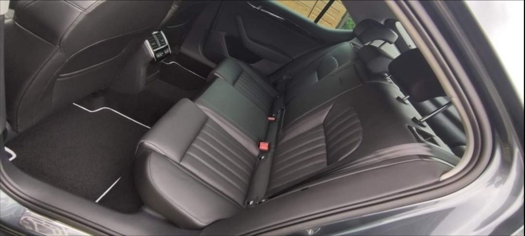 Wnętrze samochodu osobowego z czarną skórzaną tapicerką, widok na tylną kanapę z pasami bezpieczeństwa i dywaniki z białymi lamówkami.