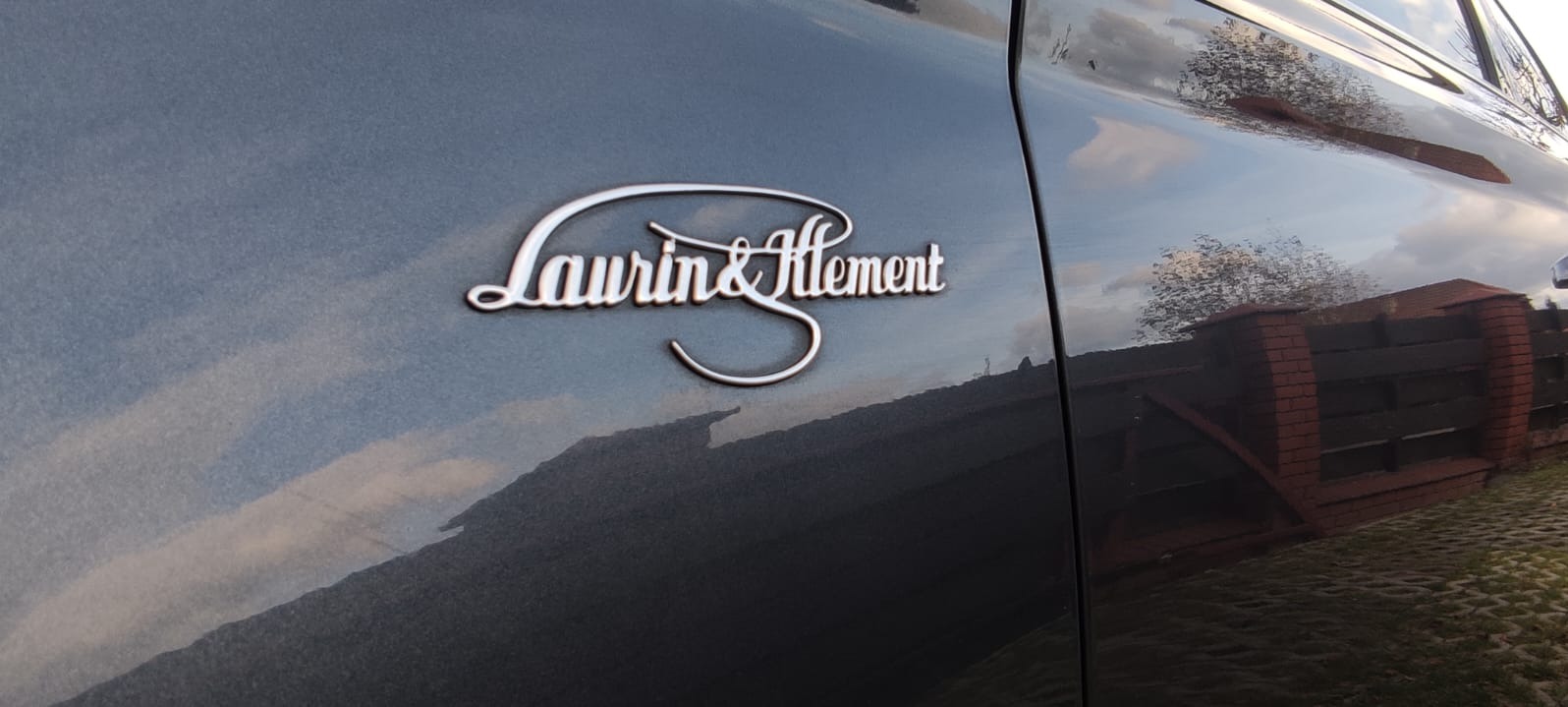 Chromowany napis Laurin&Klement na boku grafitowego samochodu z odbiciem nieba i drzewa
