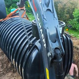 Hydro-Term - Ciężki sprzęt marki Schaeff opuszcza czarny, żebrowany zbiornik na szambo do wykopu, widoczny pracownik w dole.