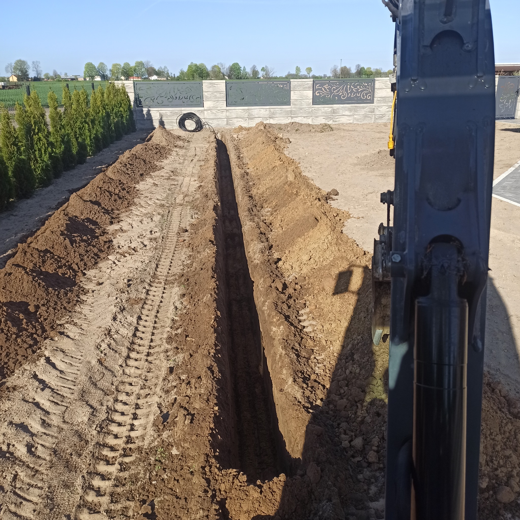 Długi, wąski rów wykopany w ziemi, widoczne ślady opon koparki, w tle ozdobny betonowy płot i równe szpalery tuj.