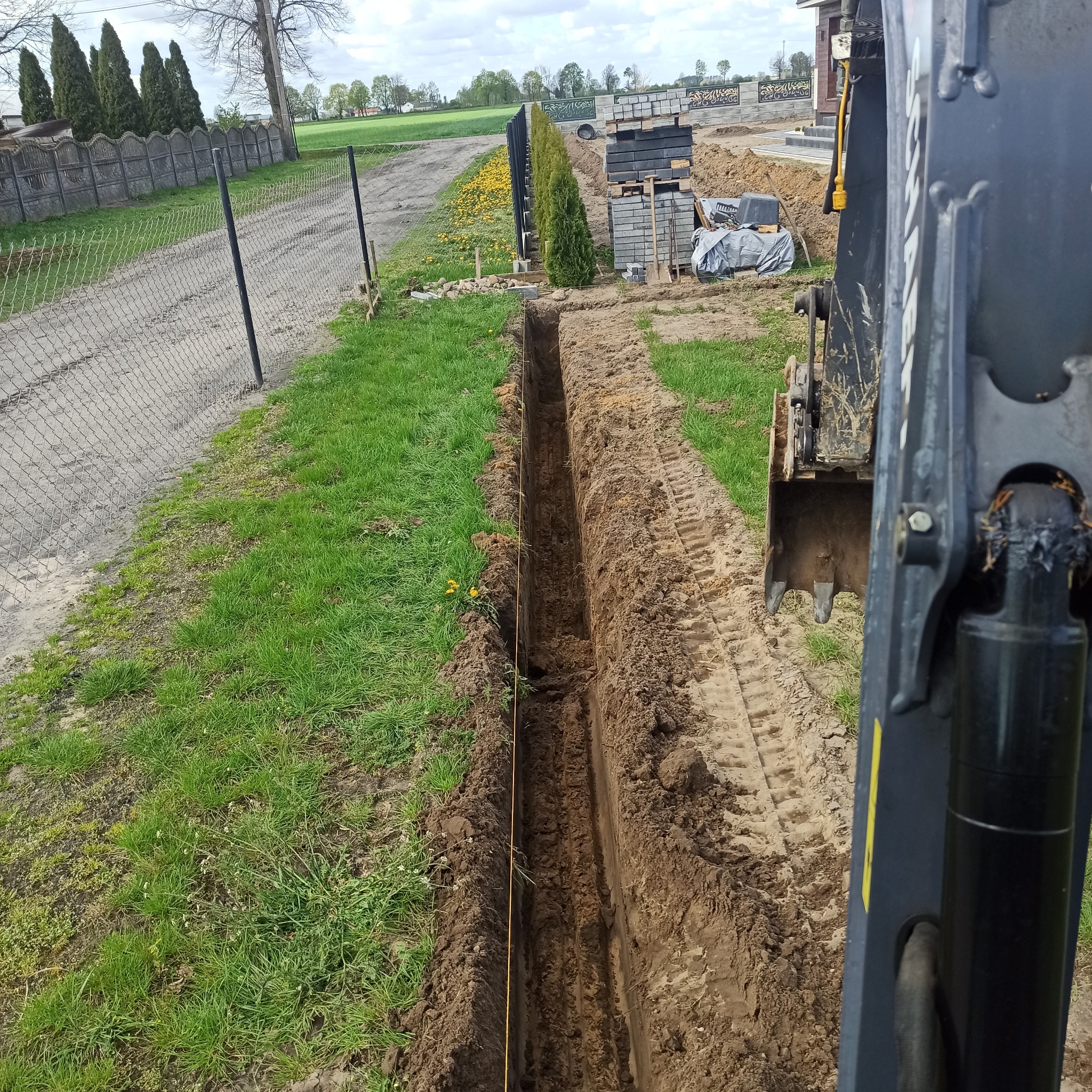 Długi, wąski wykop w ziemi, wyznaczony sznurkiem, z boku widoczna część koparki i ułożone bloczki betonowe.