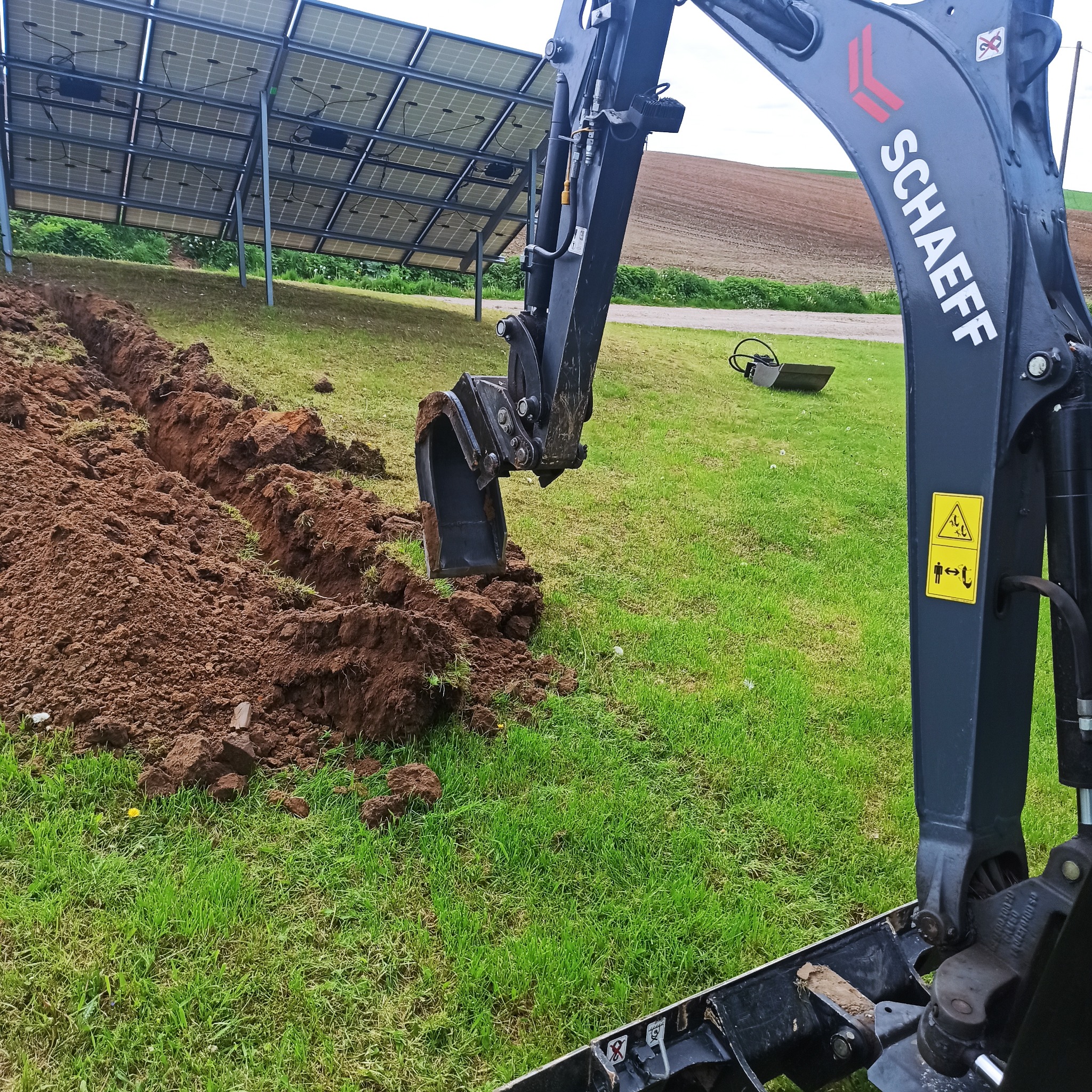 Ciężki sprzęt marki Schaeff wykopuje rów na trawiastym terenie w pobliżu paneli słonecznych.