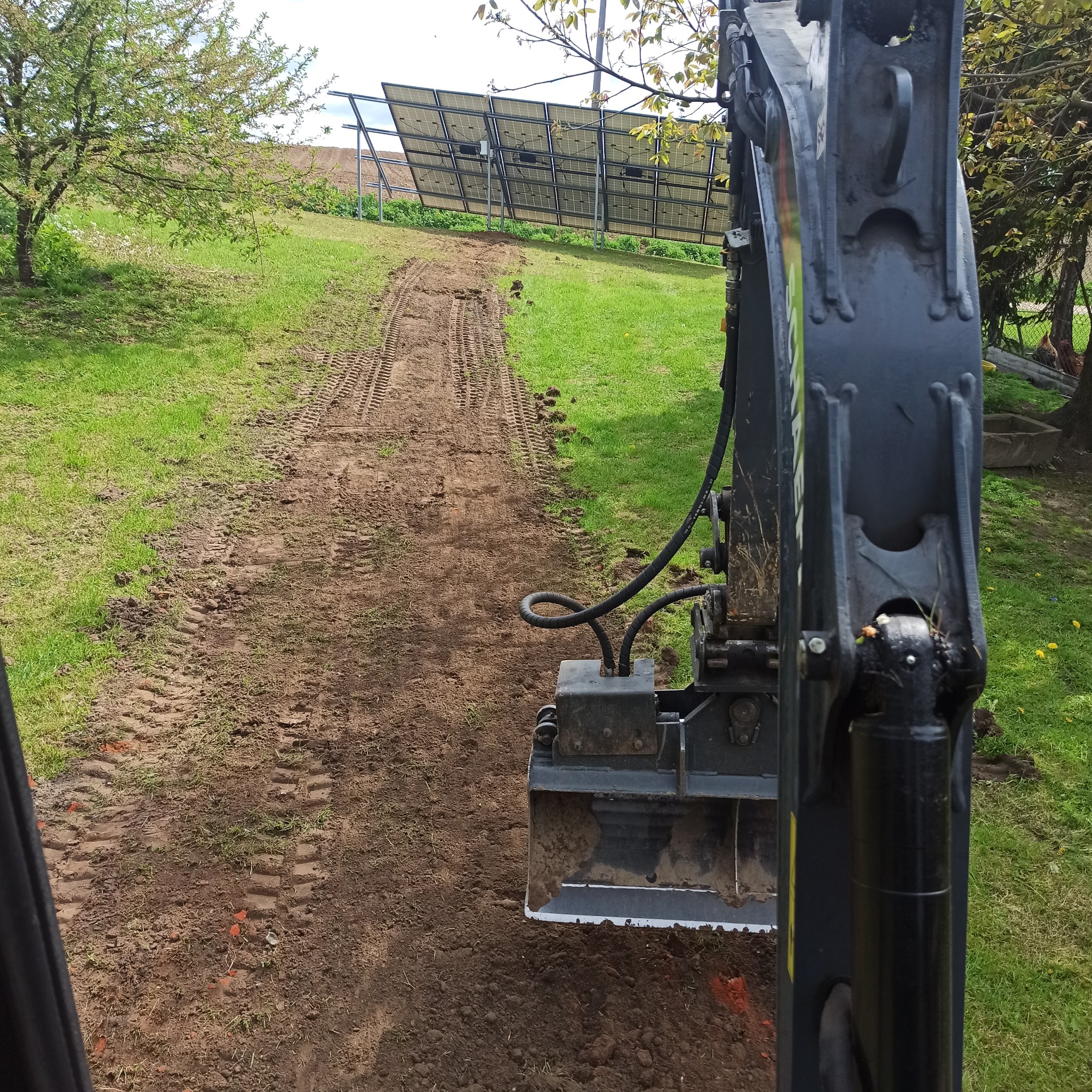 Widok z kabiny operatora na czarną maszynę do robót ziemnych, która wyrównuje teren pod panele słoneczne na wzgórzu, widoczne ślady opon na ziemi.