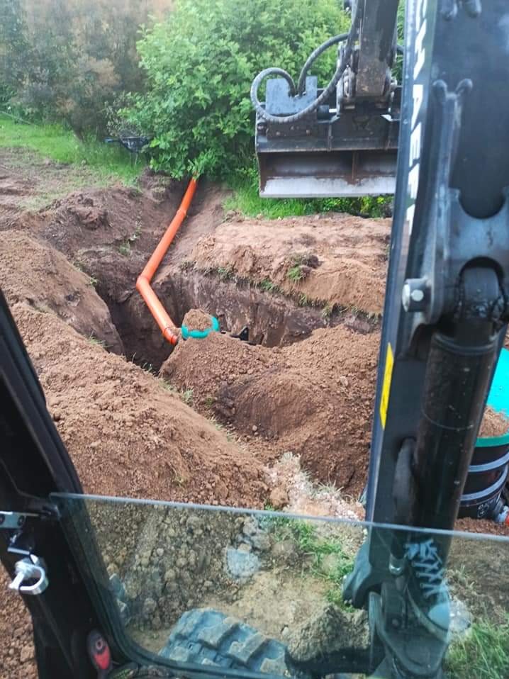 Pomarańczowe rury ułożone w wykopie ziemnym, widok z kabiny koparki, zieleń w tle.