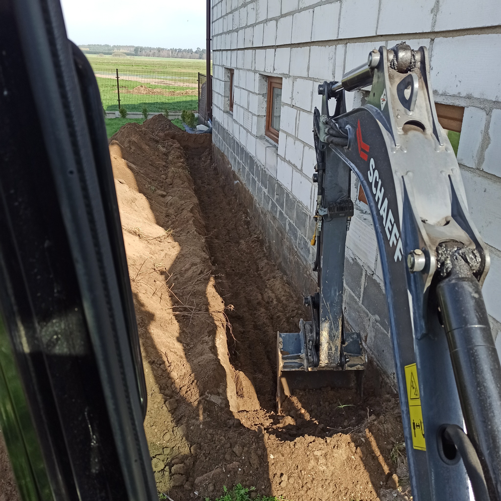 Mini koparka Schaeff wykonuje wąski wykop wzdłuż ściany budynku z białego bloczka, widok z kabiny operatora.