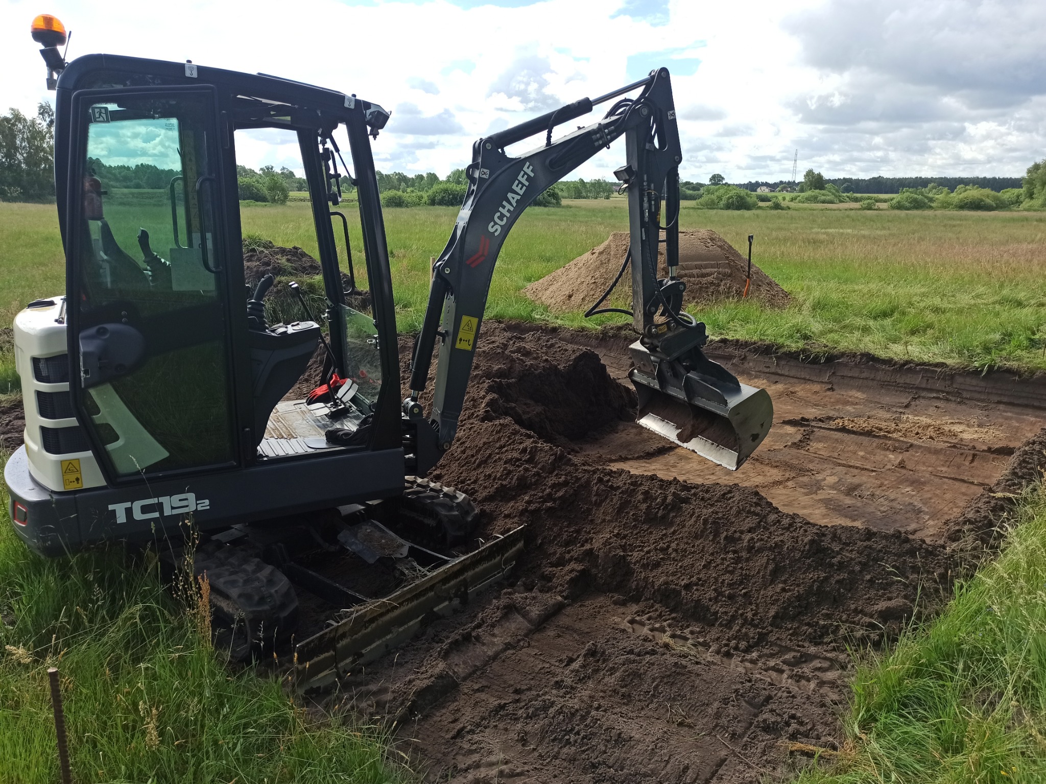 Mini koparka Schaeff TC19.2 podczas prac ziemnych na zewnątrz, widoczne wykopane zagłębienie w ziemi i usypana pryzma ziemi w tle, w oddali łąka i drzewa.