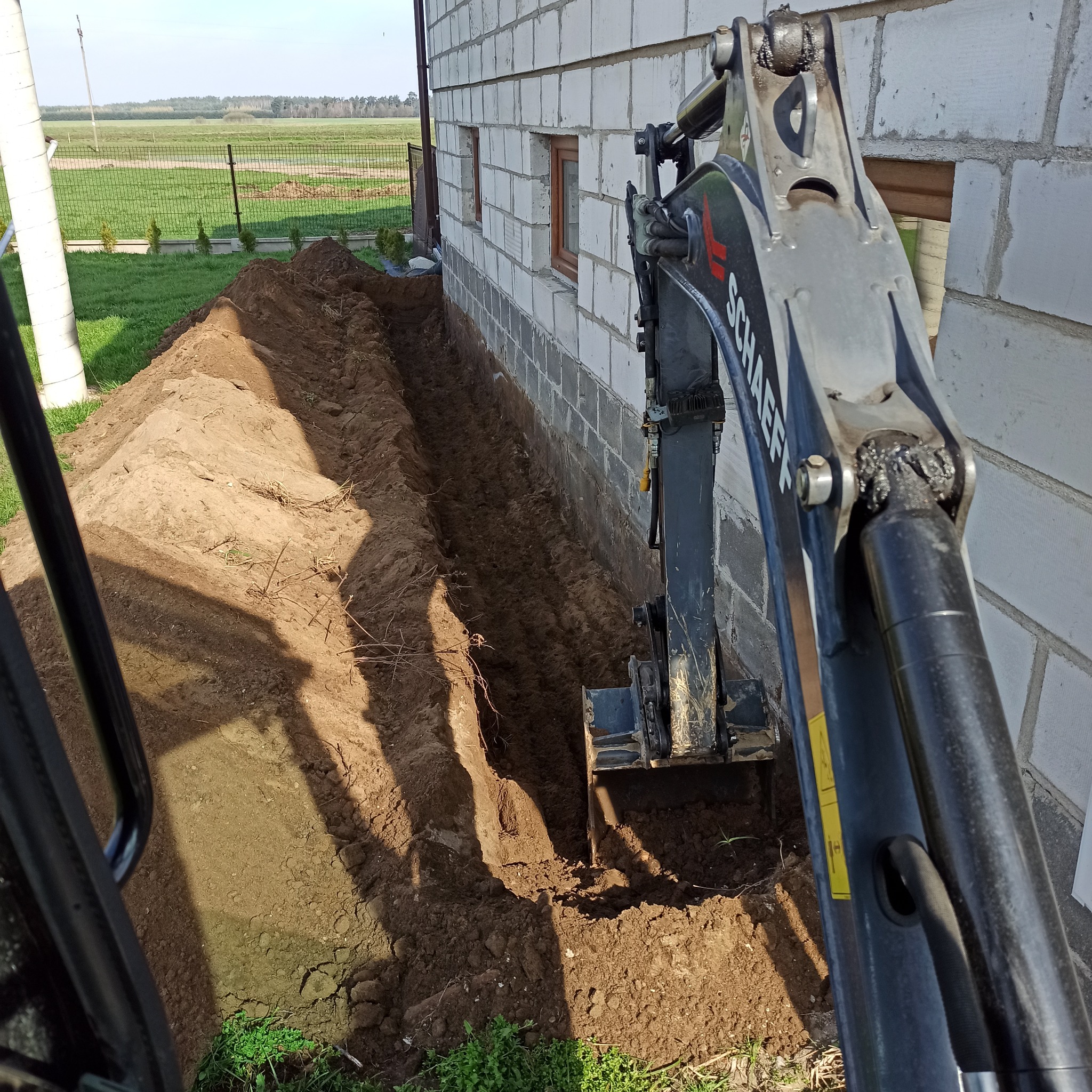 Mini koparka Schäeff wykopuje wąski rów wzdłuż ściany budynku z białego pustaka, z hałdą ziemi po jednej stronie rowu, widok z kabiny operatora.