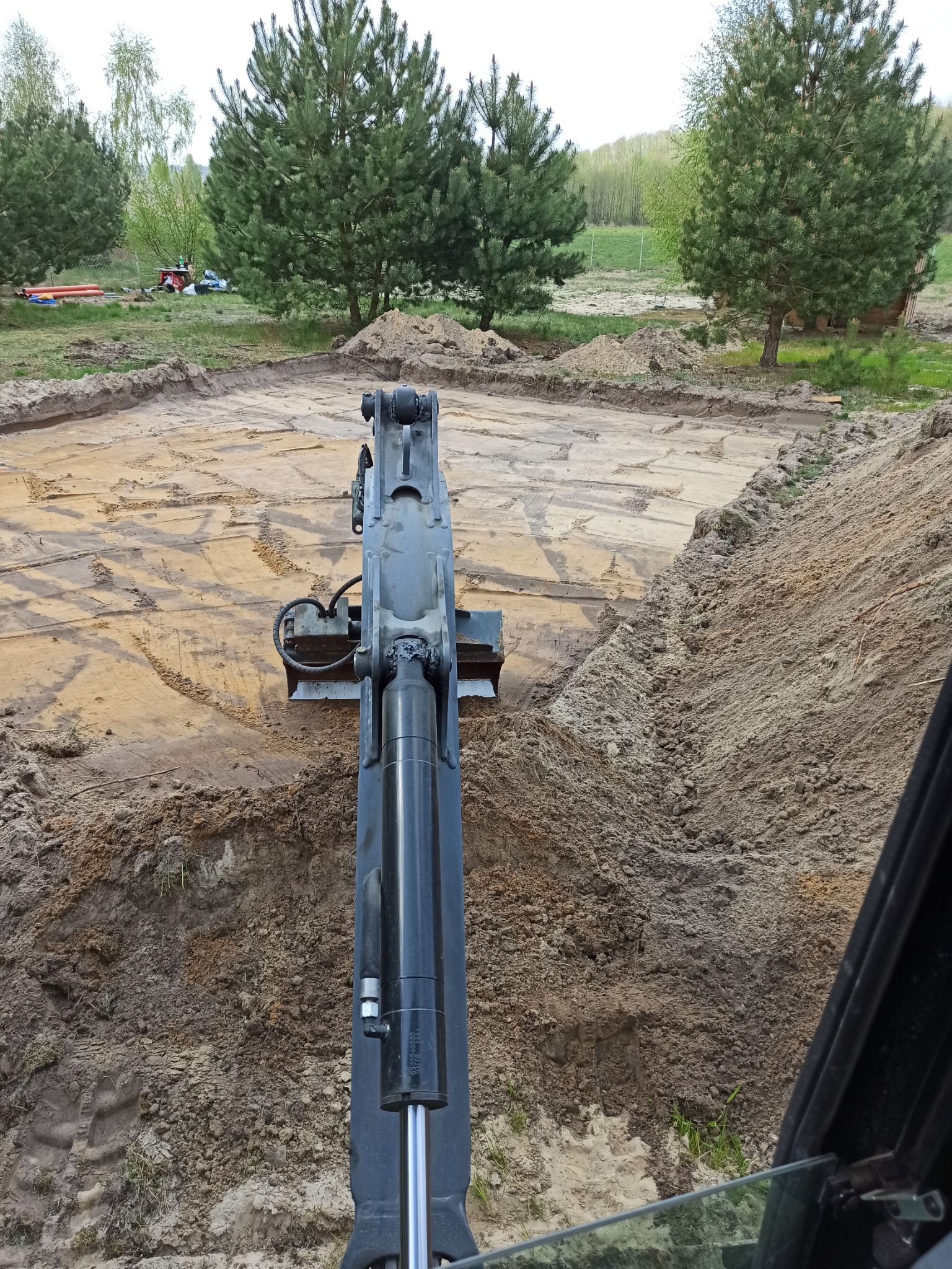 Widok z kabiny koparki na wykop pod fundamenty, widoczne ślady pracy łyżki, w tle drzewa i sprzęt budowlany.