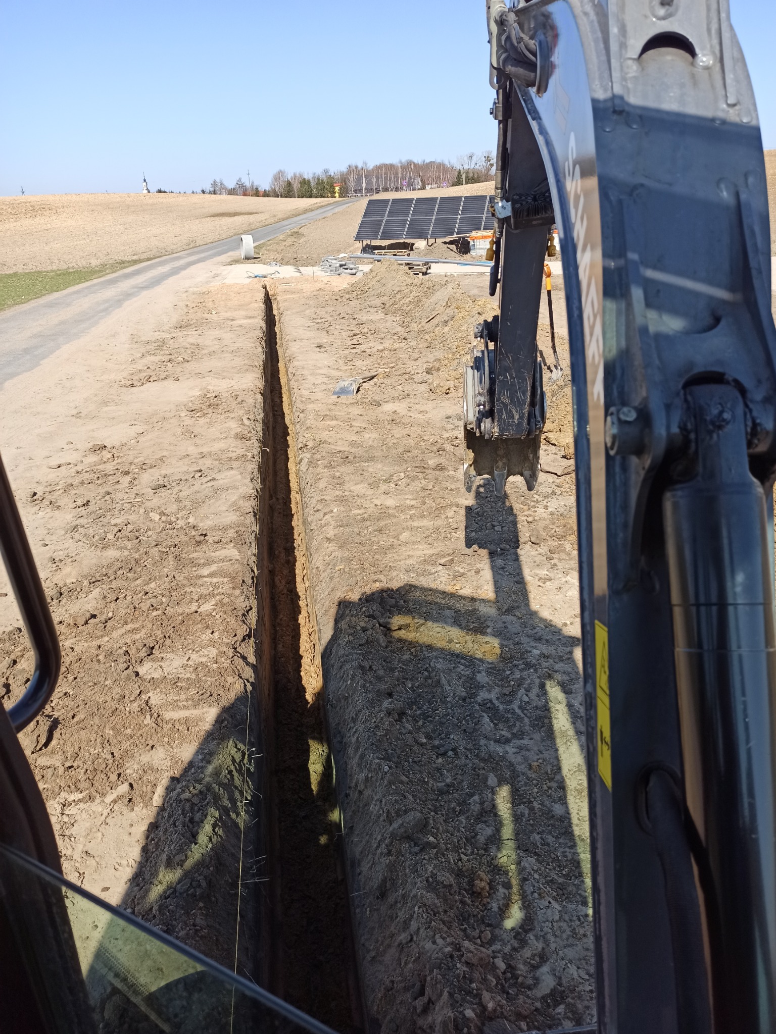 Wąski wykop w piaszczystej ziemi, widok z kabiny koparki na tle paneli słonecznych i odległego kościoła.