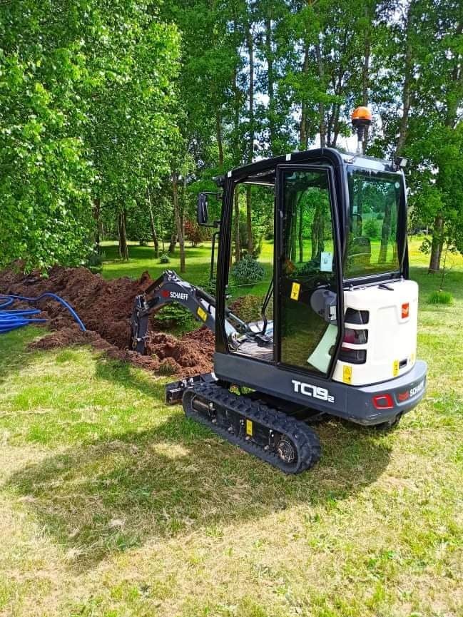 Mini koparka Schaeff TC192 wykonuje wykop w trawiastym terenie, widoczne niebieskie rury i świeżo wykopana ziemia.