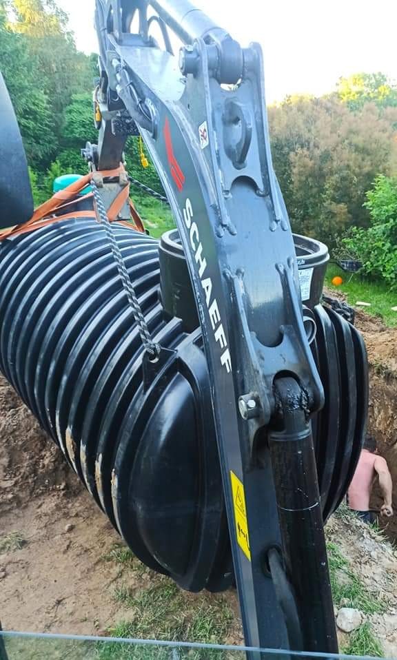 Ciężki sprzęt marki Schaeff opuszcza czarny, żebrowany zbiornik na szambo do wykopu, widoczny pracownik w dole.