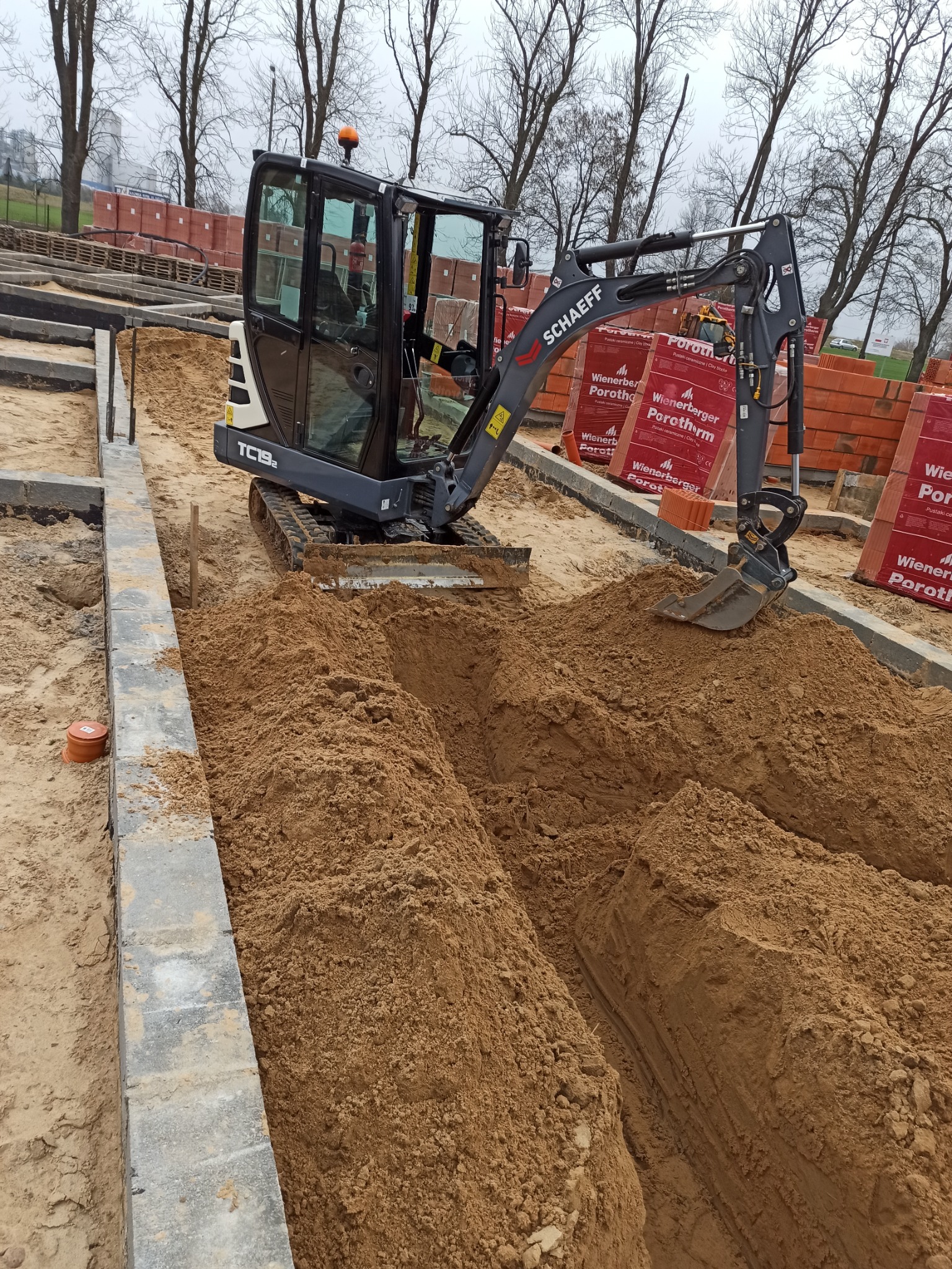 Mała koparka Schaeff TC192 wykonuje wąski wykop w piaszczystej glebie, obok ułożone bloczki fundamentowe i palety z cegłami Porotherm, widoczne drzewa bez liści w tle.