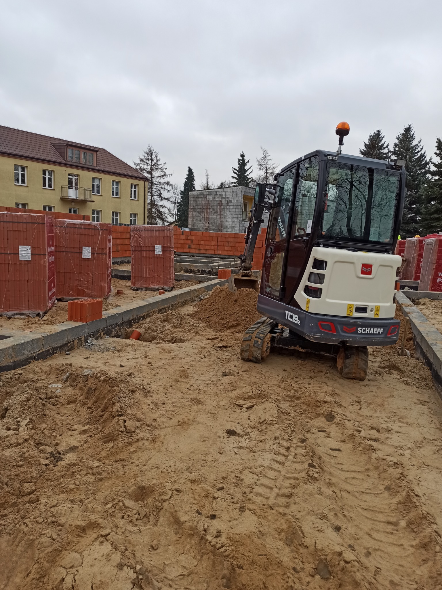 Mini koparka Schaeff TC19e na placu budowy, widoczne wykopane fundamenty, cegły paletowane w tle, pochmurne niebo.