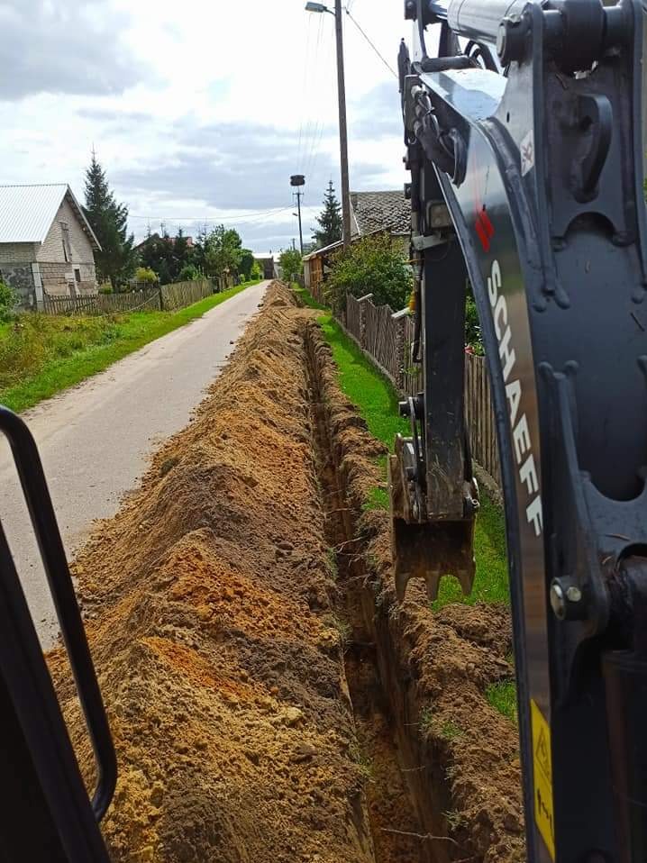 Długi, wąski wykop wzdłuż drogi wiejskiej, widoczny z perspektywy operatora koparki Schaeff, z domami i zielenią w tle.