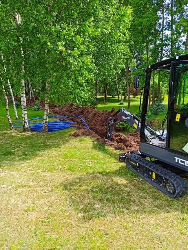 Mała koparka Schaeff wykopuje wąski rów na trawniku w pobliżu drzew, w rowie ułożone niebieskie rury.