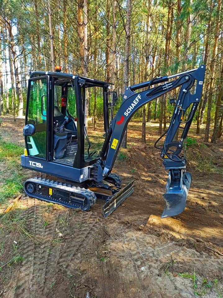 Niewielka, czarno-szara koparka Schaeff TC19-2 z uniesioną łyżką stoi na ziemi w lesie sosnowym, widoczne ślady opon na piasku.