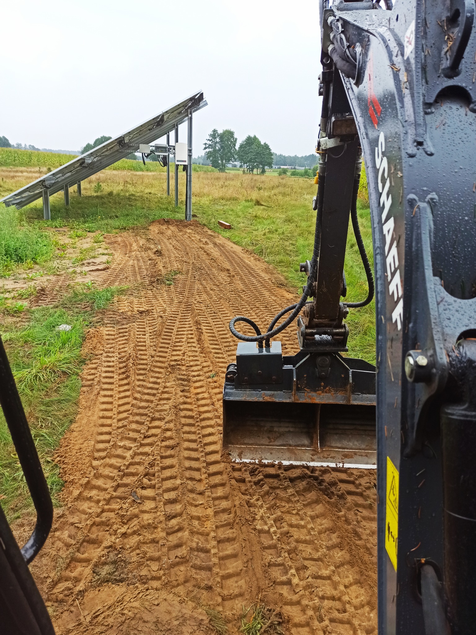 Ciężka koparka Schaeff wykopuje rów pod instalację paneli słonecznych na lekko pochylonym, trawiastym terenie, widok z kabiny operatora.