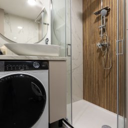 NIFU Jeremi Bombała - Nowoczesna łazienka z kabiną prysznicową typu walk-in z deszczownicą, umywalką nablatową na szafce i pralką z czarnym okrągłym oknem, wykończona białym marmurem i drewnianymi panelami ściennymi.