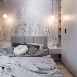 NIFU Jeremi Bombała - Nowoczesna sypialnia z tapetą w motywie liści palmowych, szarą tapicerowaną ramą łóżka, okrągłą białą poduszką dekoracyjną i wysoką, białą szafą w zabudowie z czarnymi uchwytami.